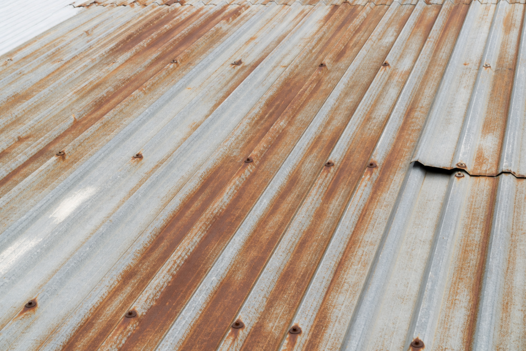 Corrugated metal roof with widespread rust streaks from fasteners, surface discoloration, and a slightly lifted panel edge.
